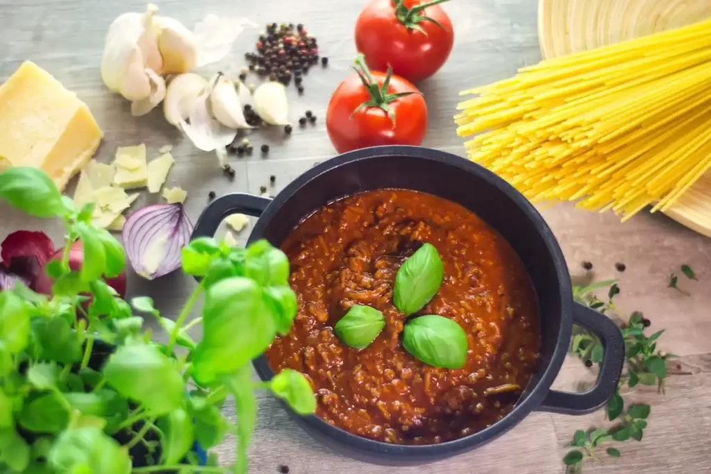 Une sauce bolognaise dans une marmite posée à côté de pâte, des herbes, des tomates et des oignons