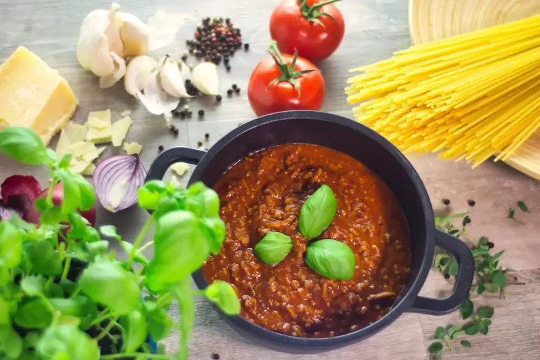 Une sauce bolognaise dans une marmite posée à côté de pâte, des herbes, des tomates et des oignons